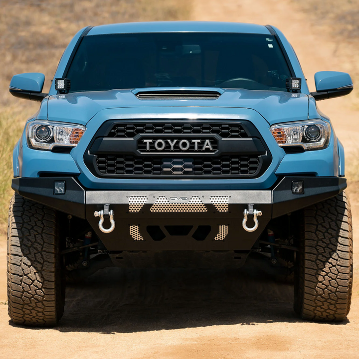 A blue Tacoma truck with a black front bumper featuring the Toyota logo, designed for off-road use, with visible tire treads and a light bar mount.