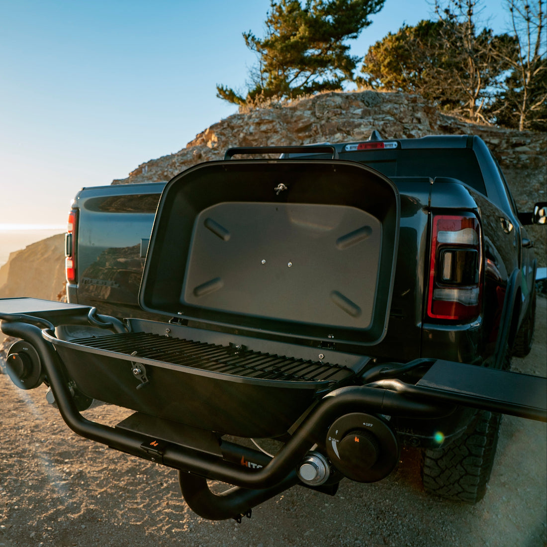 Back of a black truck with an open tailgate on a rocky beach with ocean and sunset in the background.
