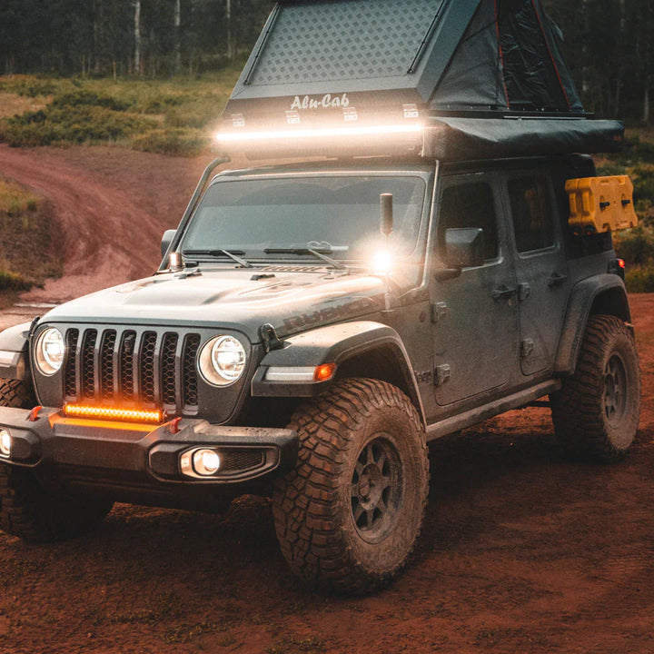Grey Jeep Wrangler Rubicon with Heretic Studio LED fog lights and a rooftop tent on a dirt road.